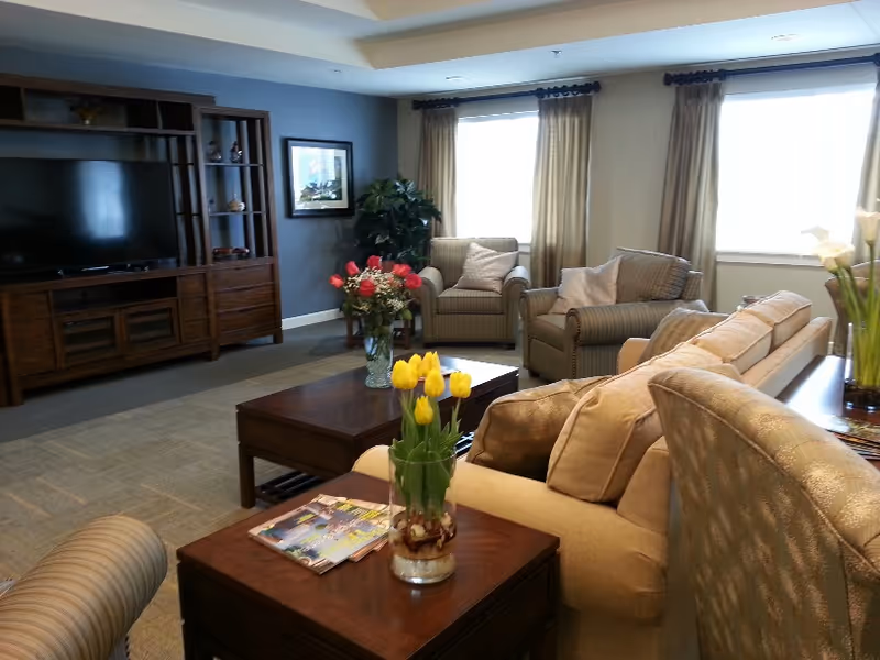 A cozy living room with beige and striped armchairs and sofas arranged around wooden coffee tables. There are vases with yellow tulips and red flowers on the tables. A large wooden entertainment center with a TV is against the wall, and two large windows with beige curtains let in natural light. A framed picture and a green plant are also visible in the room.