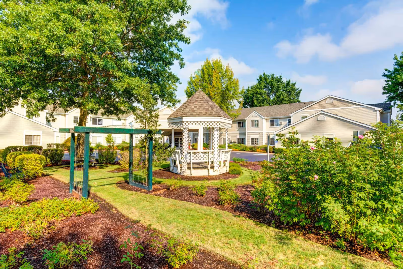 A bright outdoor garden area at The Oaks at Lebanon featuring a white gazebo with lattice details, surrounded by green bushes, trees, and well-maintained landscaping. Beige residential buildings are visible in the background under a blue sky with scattered clouds.