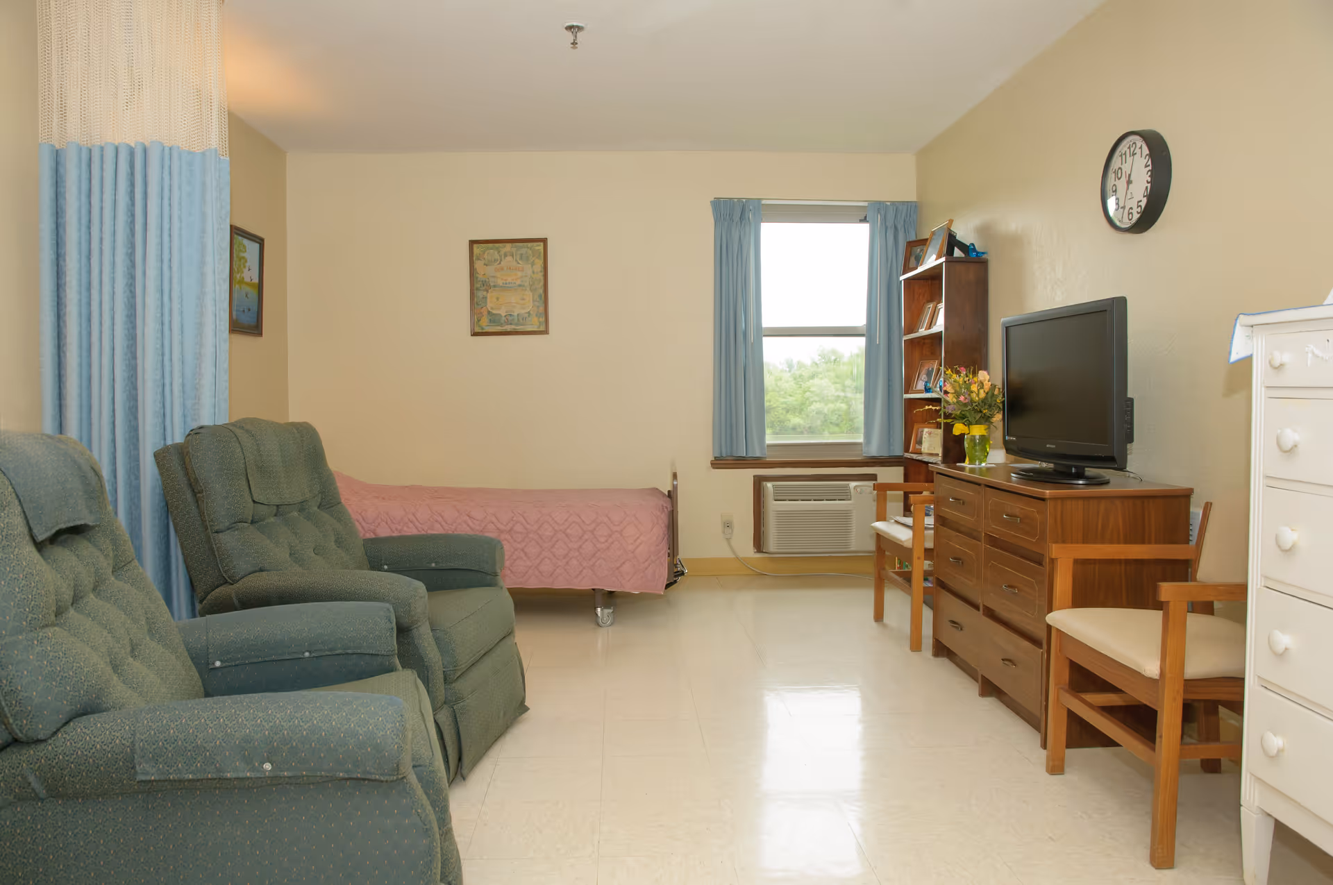 A senior living facility room with two green upholstered armchairs, a single bed with a pink bedspread, a wooden dresser with a flat-screen TV, a wooden bookshelf, a window with blue curtains, and a wall clock.