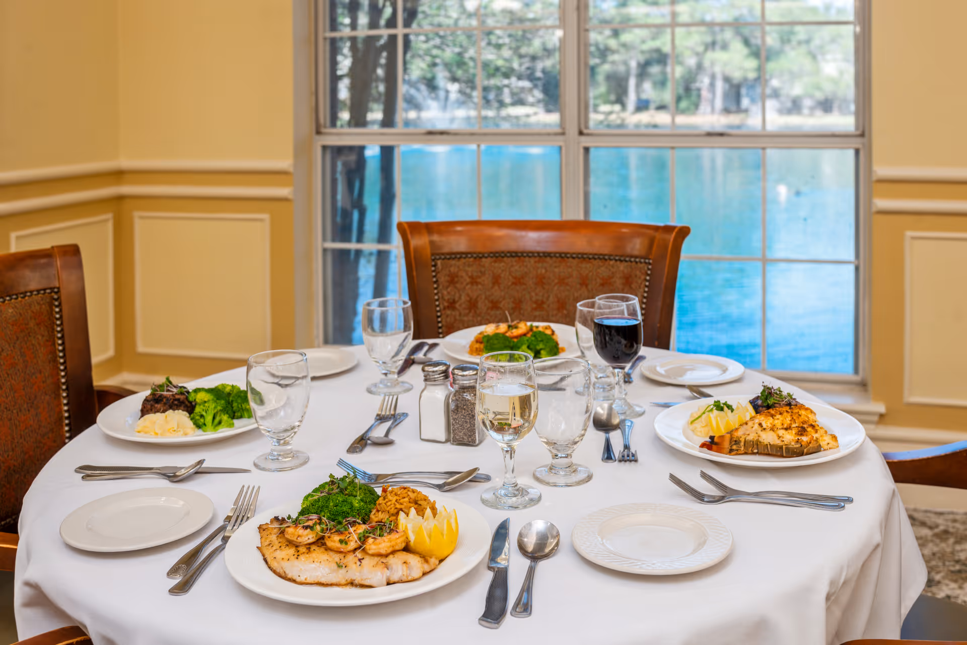 A round dining table set for four with plates of food including grilled fish, shrimp, broccoli, mashed potatoes, and rice. There are glasses of water, white wine, and red wine on the table. The table is covered with a white tablecloth and is positioned in front of a large window overlooking a body of water and greenery outside.
