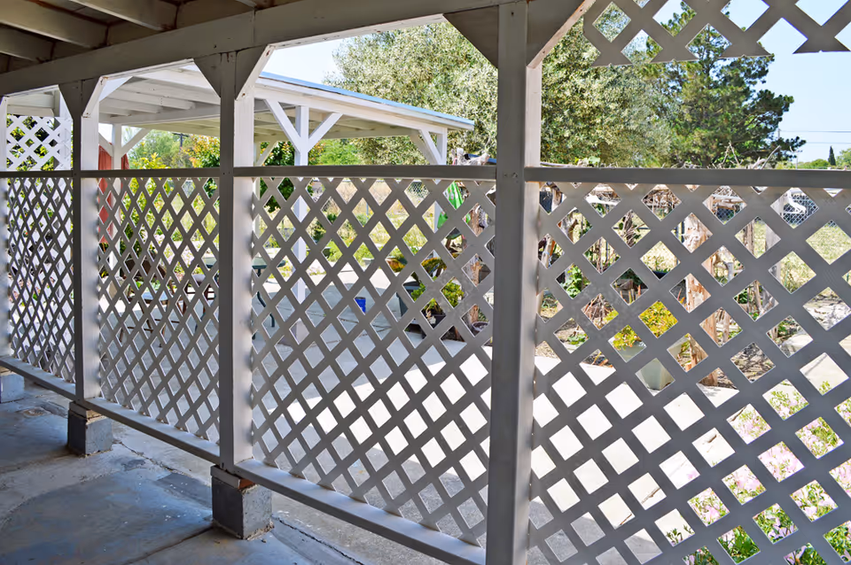 View through white lattice fencing of a covered patio area with concrete floor, some potted plants, and trees in the background under a clear blue sky.