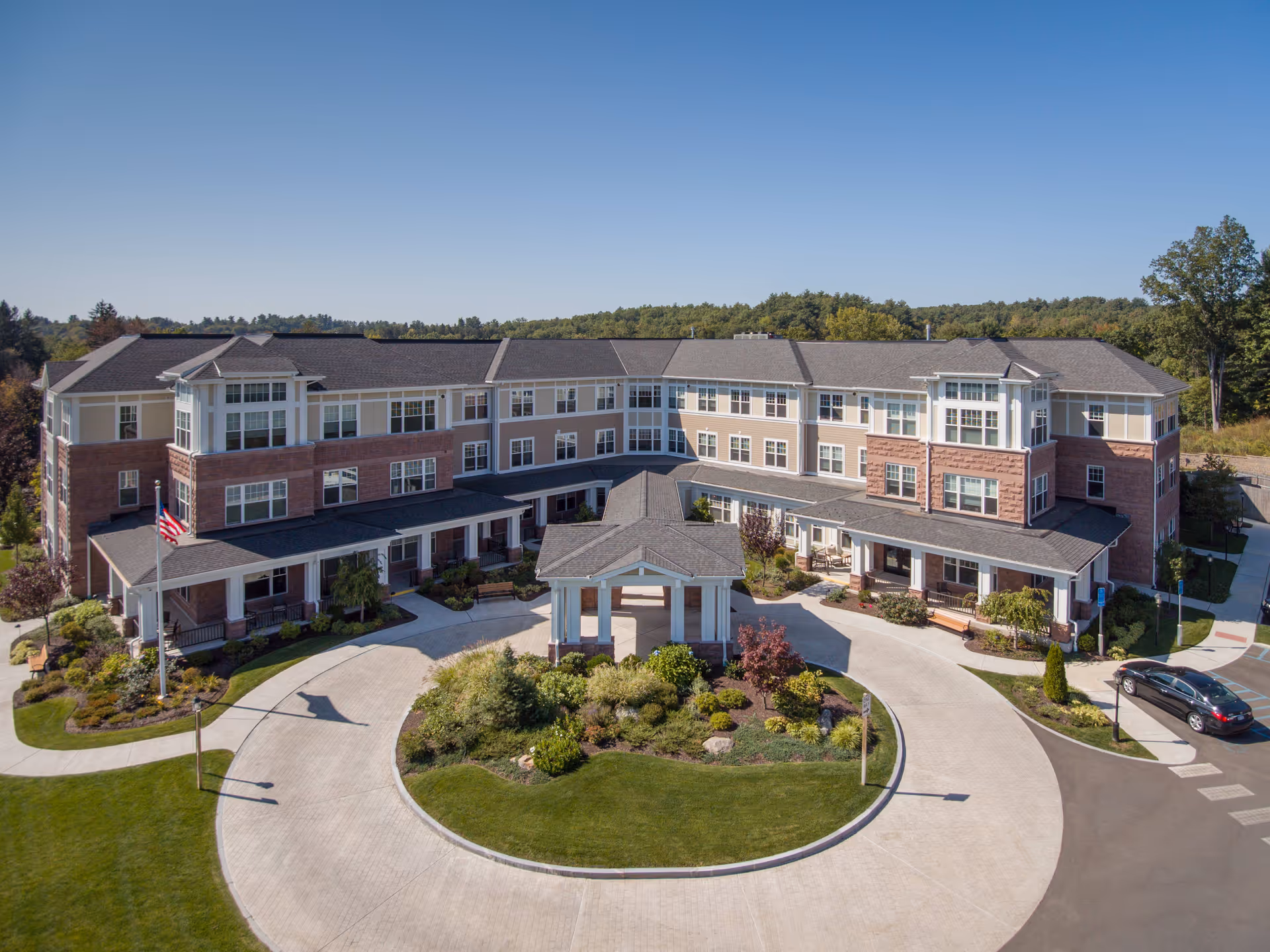 Front exterior view of a large, three-story senior living facility named The Residence at Brookside, featuring a circular driveway with landscaped greenery in the center, an American flag on a flagpole, and surrounded by trees under a clear blue sky.
