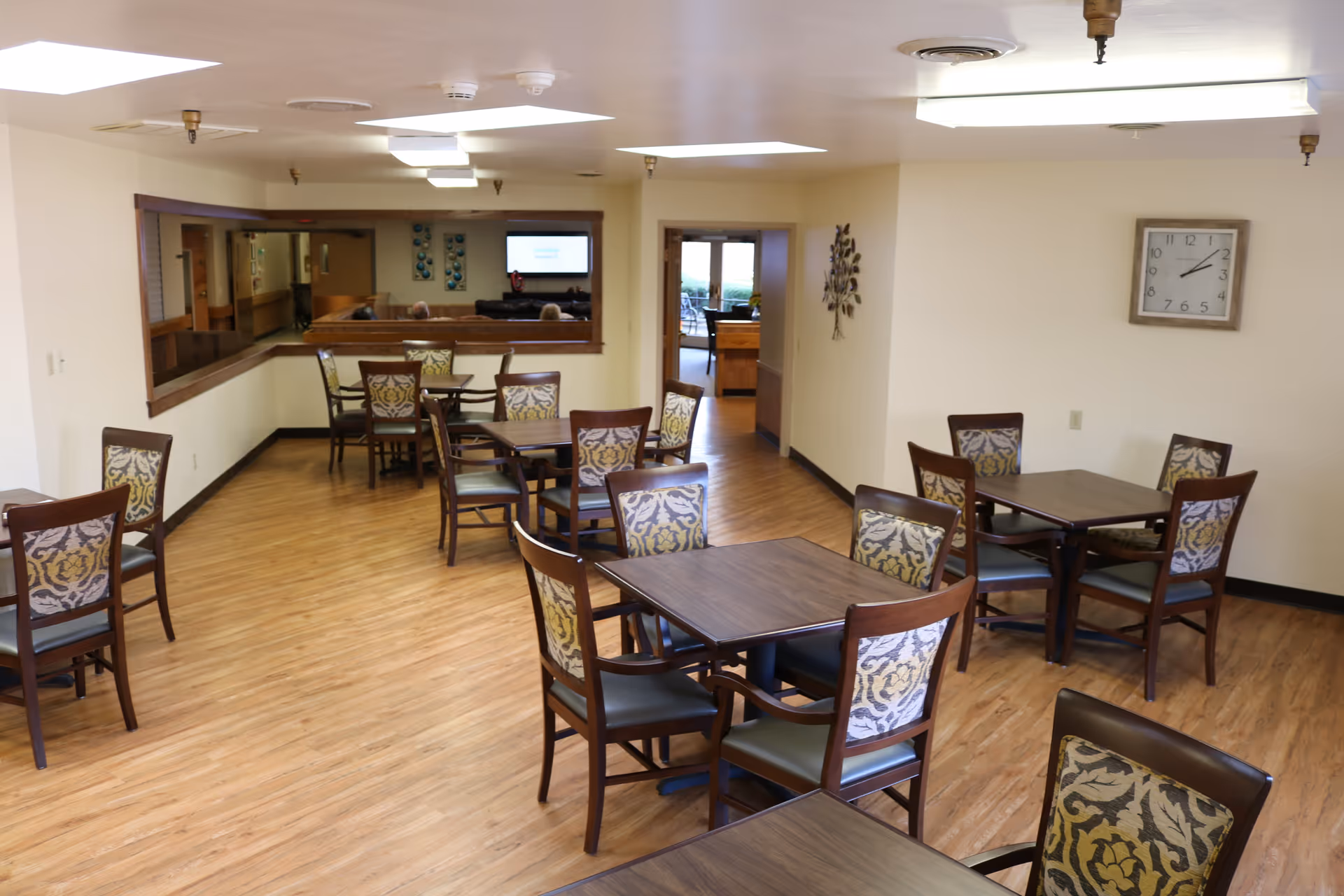 A clean and well-lit dining room with several square wooden tables, each surrounded by four chairs with patterned upholstery. The room has wood flooring, cream-colored walls, a large wall clock, and a decorative metal wall art piece. There is a large window opening to an adjacent room and a doorway leading to another area with more seating visible.