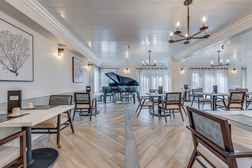 A bright and spacious dining room in Serra Highlands Senior Living featuring multiple tables and chairs arranged neatly. The room has light-colored walls adorned with framed artwork, large windows with sheer curtains allowing natural light to fill the space, and a grand piano positioned near the back. Modern chandeliers hang from the ceiling, and the floor has a wood-like tile pattern.