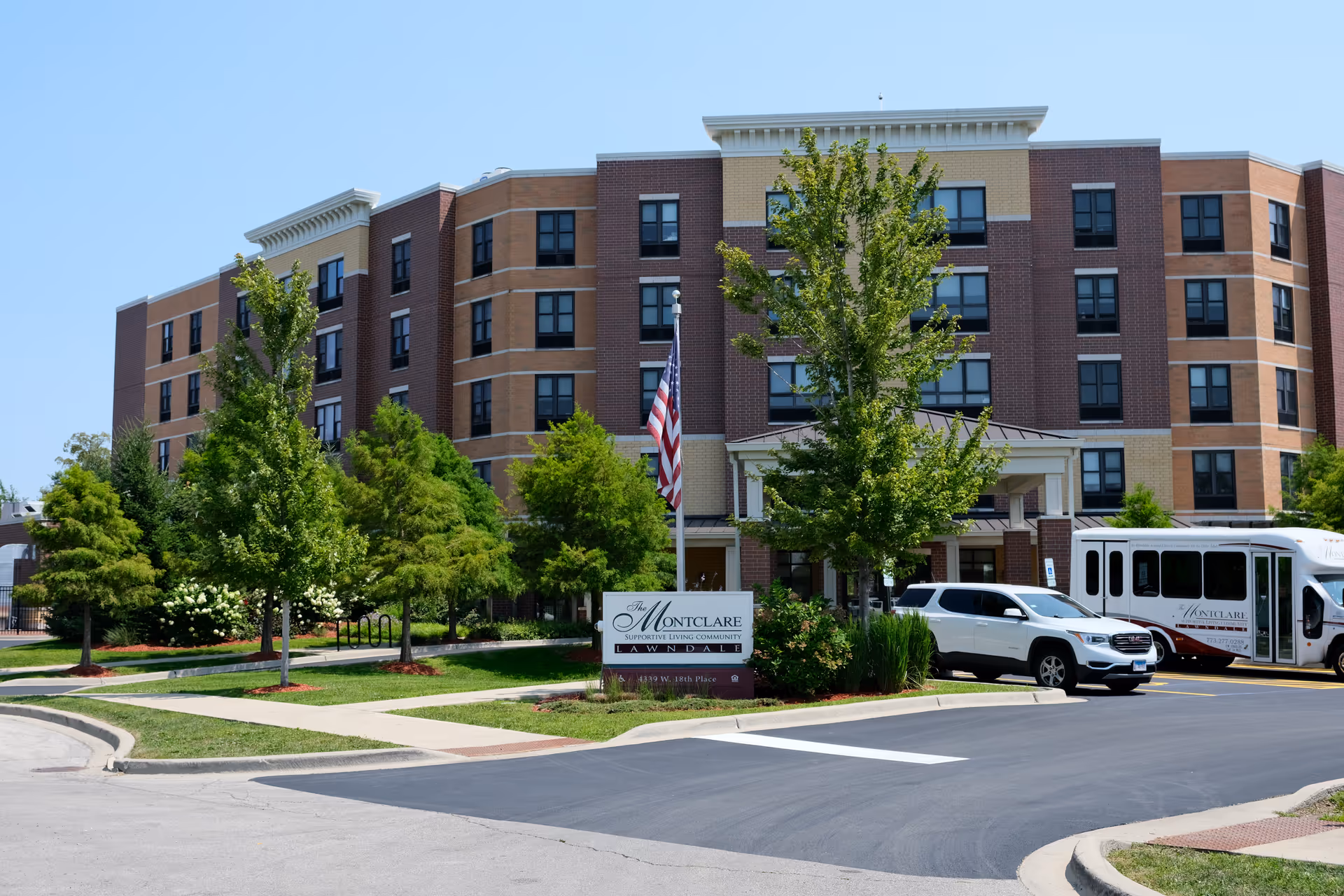 Front exterior of the Montclare Supportive Living Community building with trees, an entrance sign, a parked car and a shuttle bus.