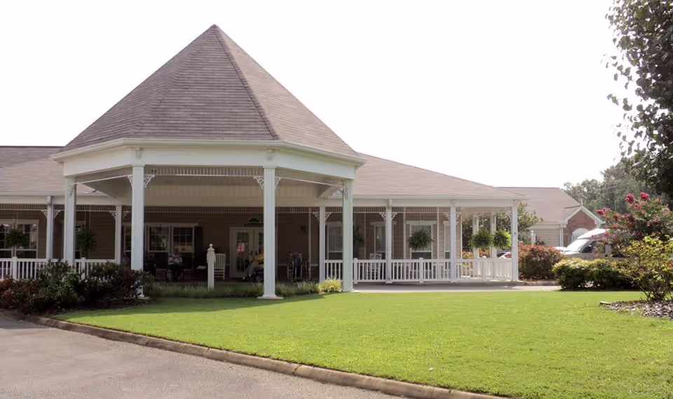 Exterior view of a senior living facility with a large covered porch supported by white columns, surrounded by green lawn and landscaping with bushes and trees.