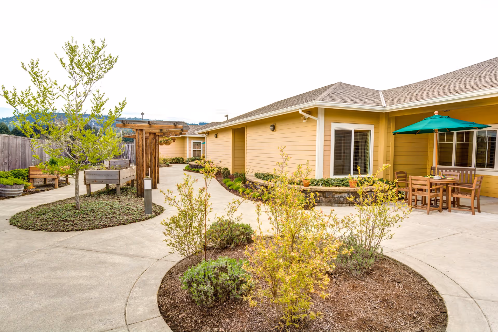 Outdoor courtyard area at Arbor Oaks Terrace Memory Care Residence featuring a curved concrete walkway, landscaped garden beds with small trees and shrubs, wooden benches, a pergola, and a patio table with chairs and a green umbrella next to a yellow building.