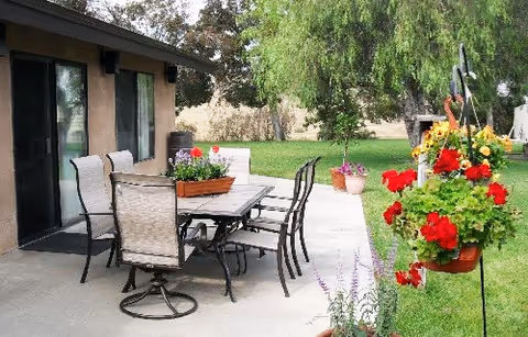Outdoor patio area with a rectangular table surrounded by six chairs on a concrete surface next to a building. There are hanging flower pots with red and yellow flowers, potted plants on the ground, and a green lawn with trees in the background.