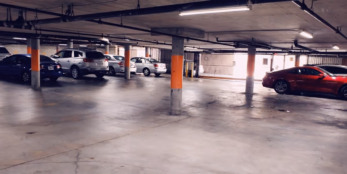Indoor parking garage with several parked cars including a red sports car and other sedans. Concrete pillars with orange markings support the ceiling, and fluorescent lights illuminate the space.