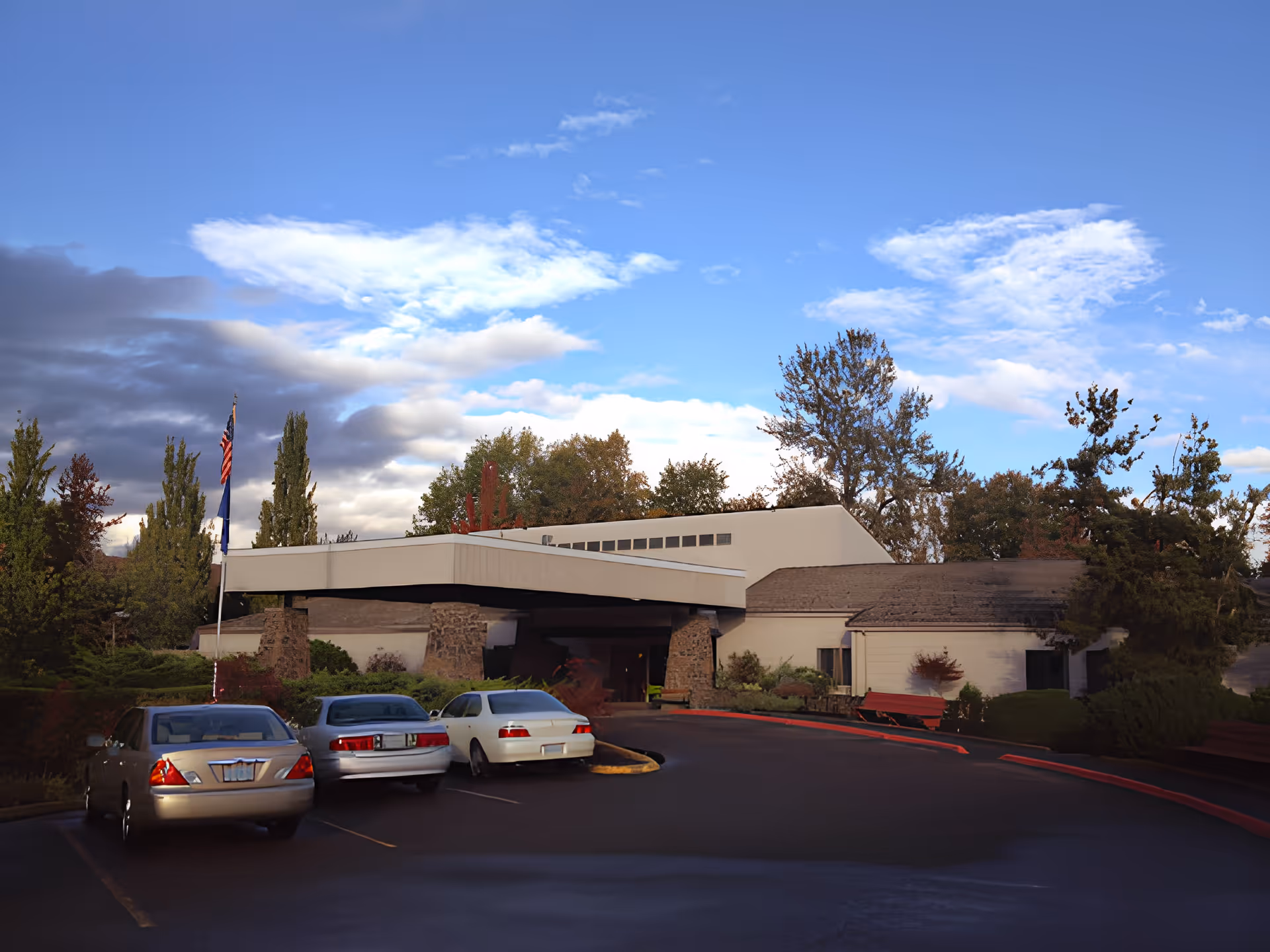 Exterior view of Churchill Estates Retirement Community building with a covered entrance supported by stone pillars, surrounded by trees and bushes. Three cars are parked in front under a partly cloudy blue sky.