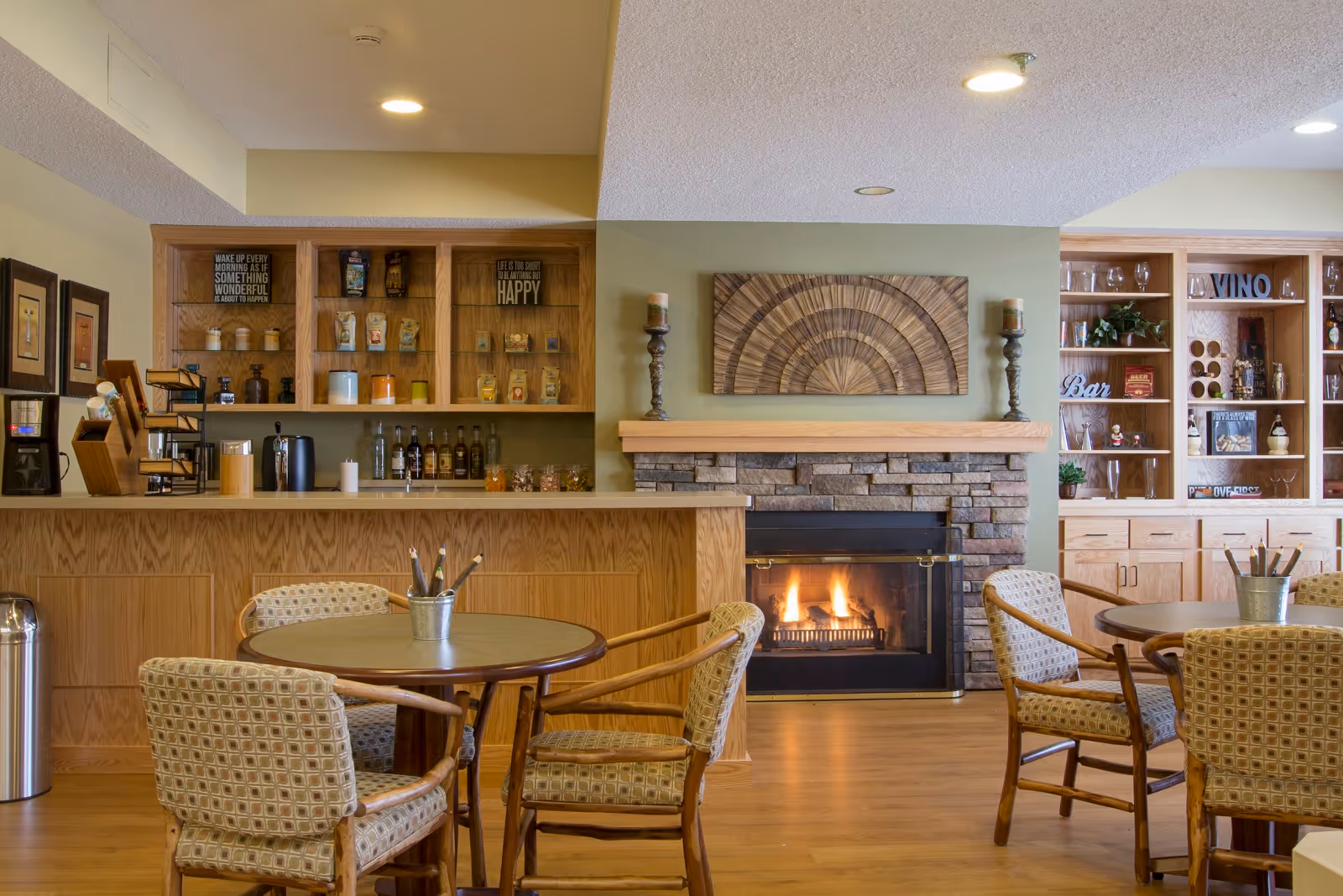 Cozy senior living common area with a stone fireplace, wooden mantel, and decorative wall art above it. There are round tables with cushioned chairs arranged around the room. Behind the tables, there is a wooden counter with shelves holding various items including bottles and decorative pieces. The room has warm lighting and a welcoming atmosphere.