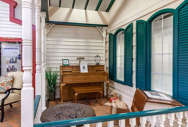 Indoor sitting area with a wooden piano against a white paneled wall, a small wooden bench in front of the piano, a decorative table with intricate metalwork, a chair with a cushion, green window shutters, and a dog statue wearing a red scarf sitting on the floor.