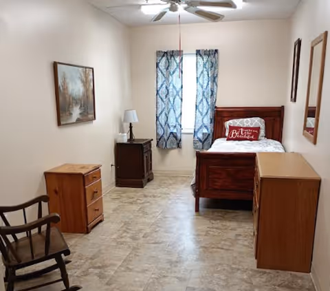 A simple, clean bedroom with a single wooden bed featuring a red pillow that says 'Be the reason someone smiles today.' The room has a wooden rocking chair, a small wooden dresser, a nightstand with a lamp, a window with blue patterned curtains, and a framed landscape painting on the wall.