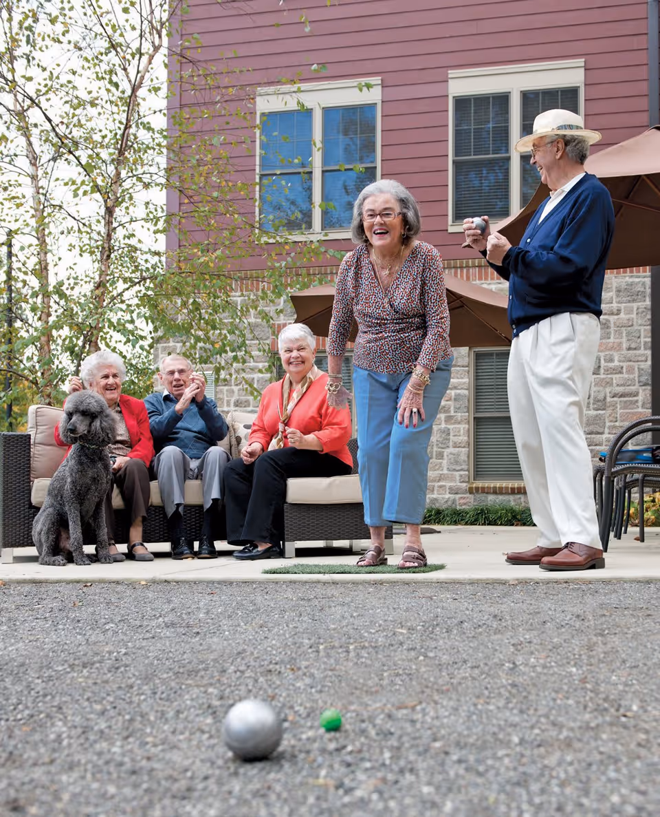 A group of elderly people enjoying an outdoor game of bocce ball on a gravel court. Three seniors are seated on a wicker outdoor sofa with a large gray dog sitting beside them, while two others stand nearby, one of whom is about to throw a bocce ball. They are in front of a building with red siding and stone accents, with trees and patio umbrellas in the background.