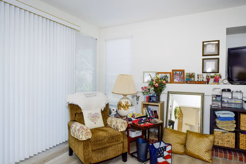 Cozy living room corner with a brown upholstered armchair adorned with a white throw and a decorative pillow. Next to the chair is a small table holding a floral lamp, books, and various items. Behind the table is a small shelving unit with flowers and framed photos on the wall above. A large mirror rests on the floor against the wall, reflecting part of the room. To the right, a TV is mounted above a shelving unit with folded towels and baskets. Vertical blinds cover the windows on the left side.