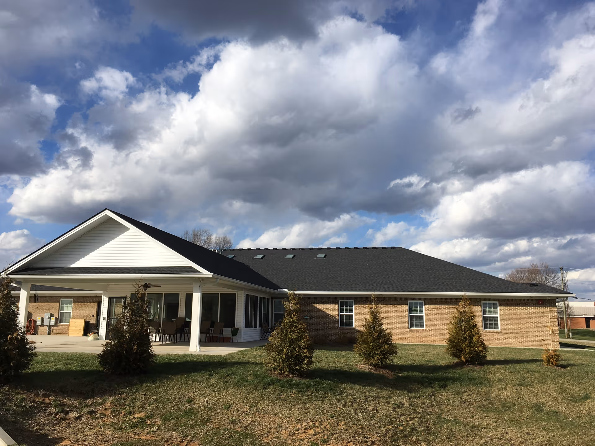 Exterior view of a single-story brick building with a dark shingled roof and white trim. The building has a covered patio area with outdoor seating and several small evergreen trees planted in the grassy yard. The sky is partly cloudy with large white and gray clouds.