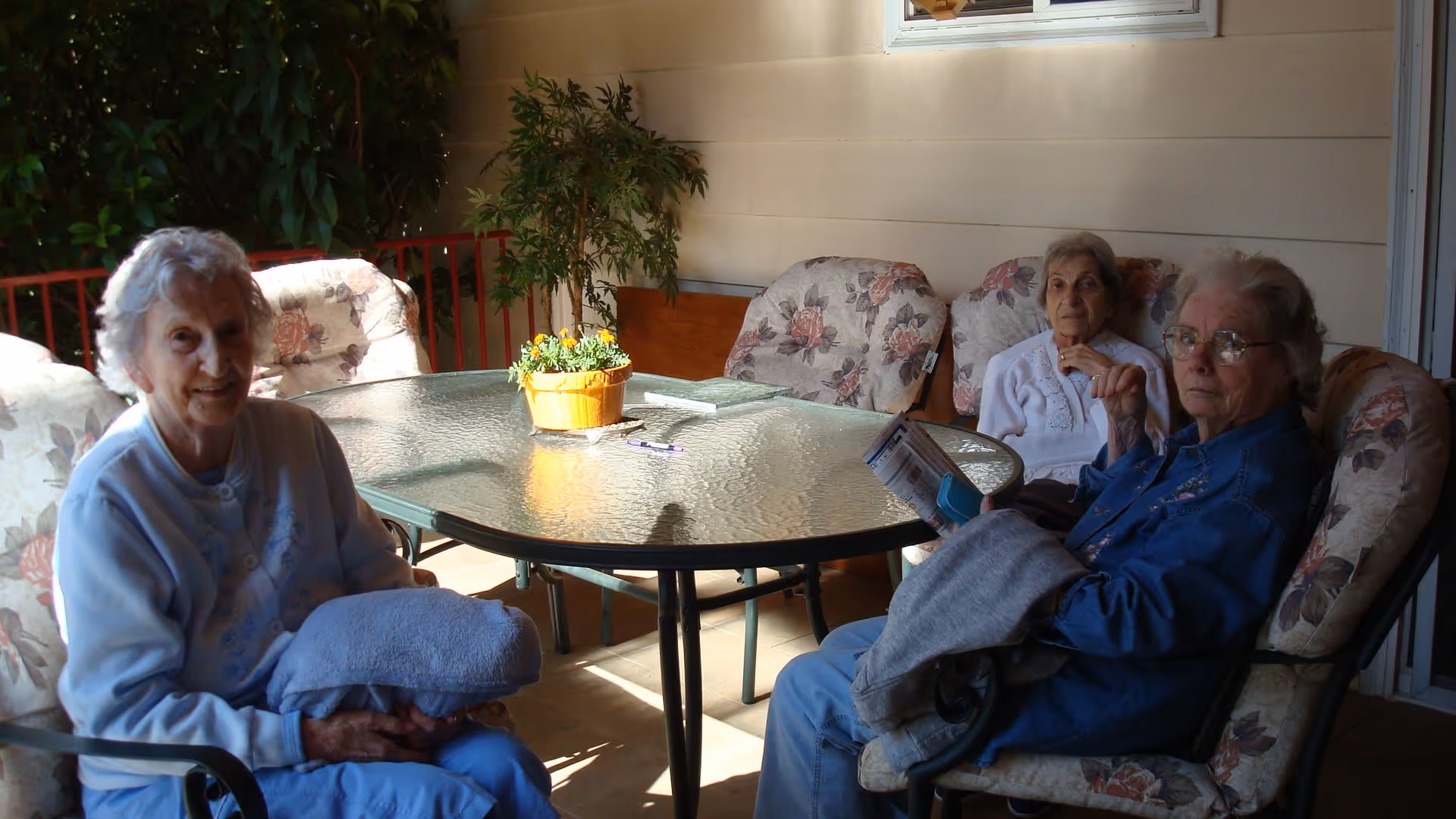 Three elderly women sitting around a glass-top outdoor table with floral cushioned chairs on a covered patio. There is a small potted plant with yellow flowers on the table and a leafy green plant in the background.