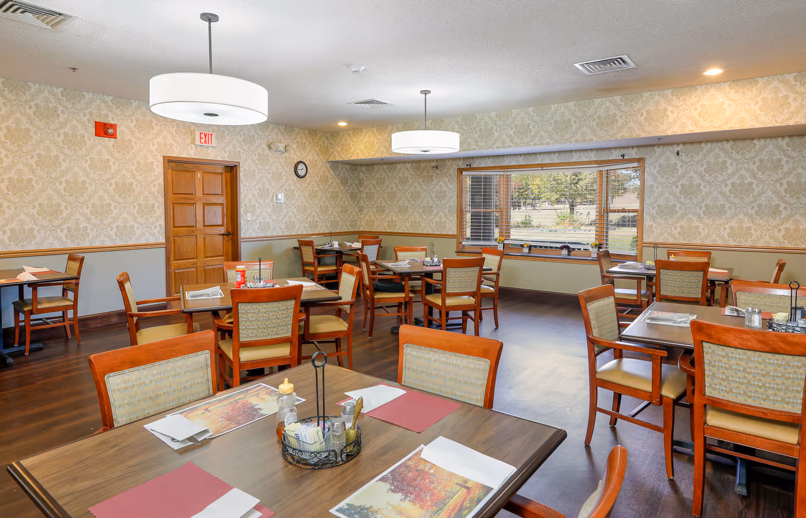 A dining room in an assisted living facility with multiple wooden tables and chairs arranged neatly. Each table has placemats, napkins, and condiments. The room has patterned wallpaper, wooden flooring, two large ceiling lights, a window with blinds showing an outdoor view, and a wooden door with an exit sign above it.