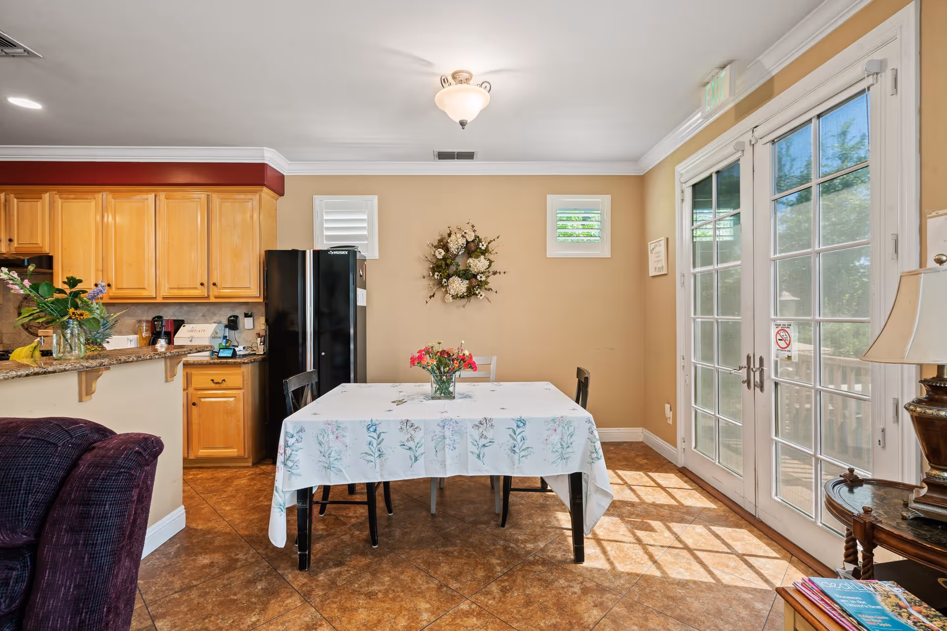 A bright dining area with a table covered by a floral tablecloth and three chairs. A vase with flowers is placed in the center of the table. To the left, there is a kitchen with wooden cabinets, a black refrigerator, and a countertop with a few items on it. On the right, there are large glass double doors letting in natural light, and a small side table with a lamp and magazines. The walls are painted beige, and there is a decorative floral wreath hanging on the wall.