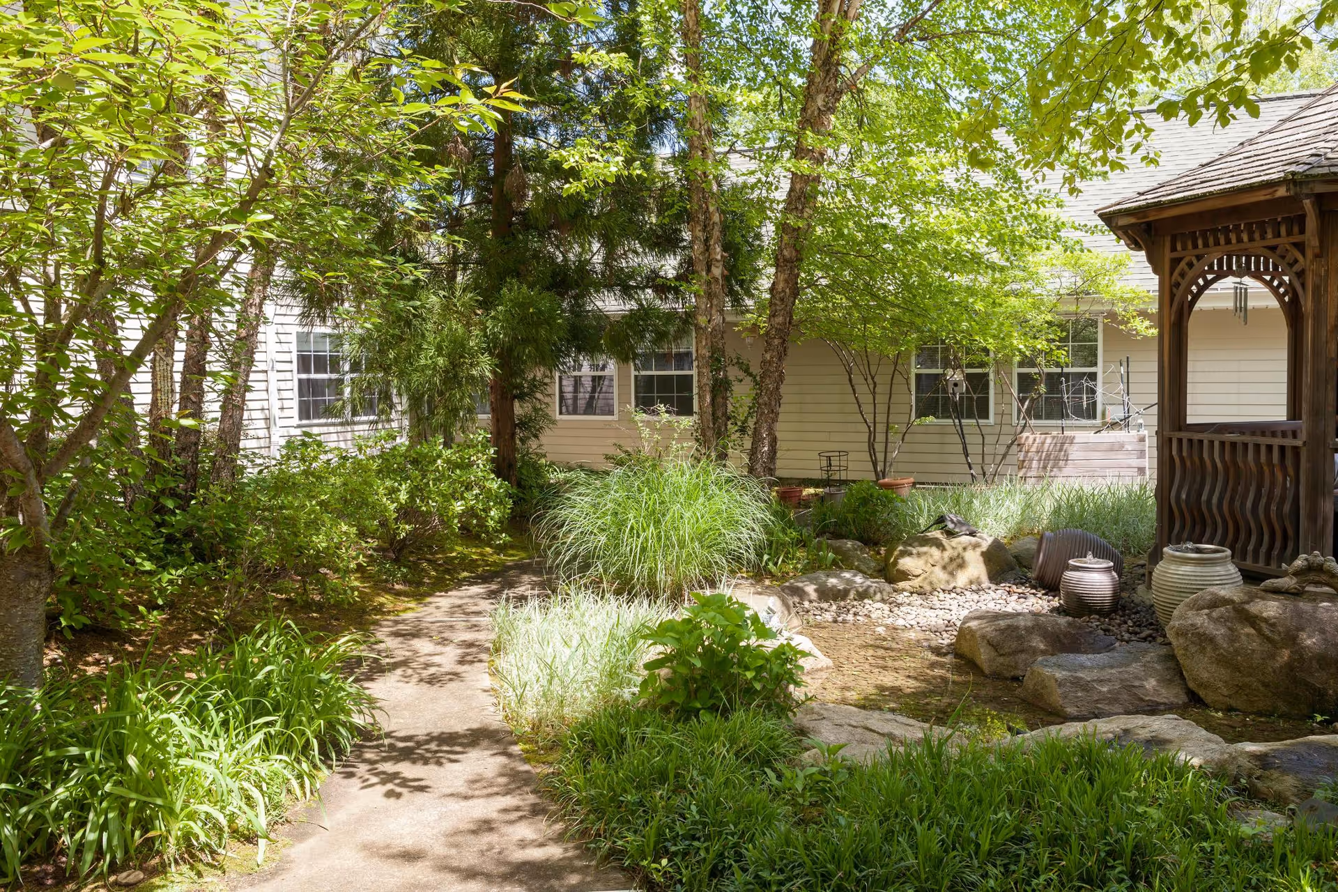 Sunlit courtyard with a winding path, lush plants, rocks, a small pond and a wooden gazebo beside a light-colored building.