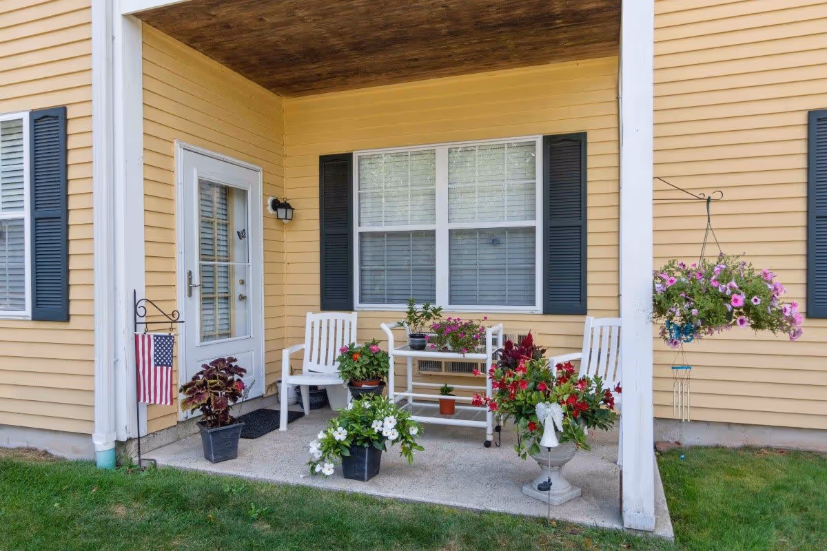 A small covered patio area attached to a yellow building with white trim and dark shutters. The patio has two white chairs, a small white table with potted plants, and several other potted flowers and plants arranged around the space. An American flag is displayed on a stand near the door, and a hanging basket with pink flowers is suspended from the patio roof. The area is surrounded by green grass.