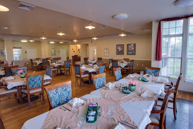 A spacious dining room with multiple tables set for meals, featuring wooden chairs with blue patterned cushions, white tablecloths with beige overlays, glassware, napkins, and small flower centerpieces. Large windows with red curtains allow natural light to fill the room, and framed artwork hangs on the beige walls.