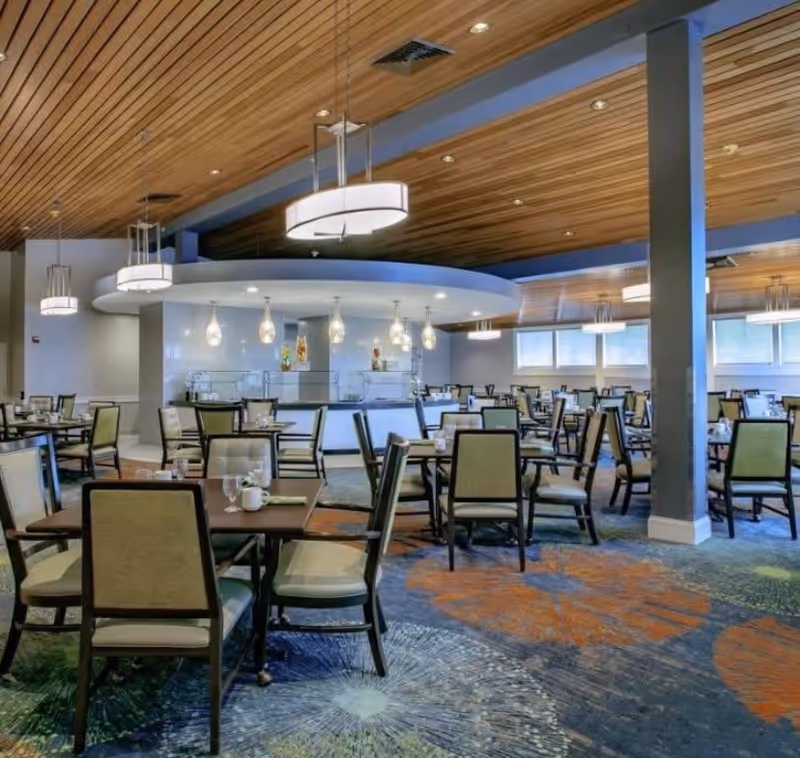 Spacious dining room with multiple tables and chairs arranged neatly. The ceiling features wooden paneling with modern hanging light fixtures. The carpet has a colorful pattern with large circular designs in orange and green. Large windows allow natural light to brighten the room.