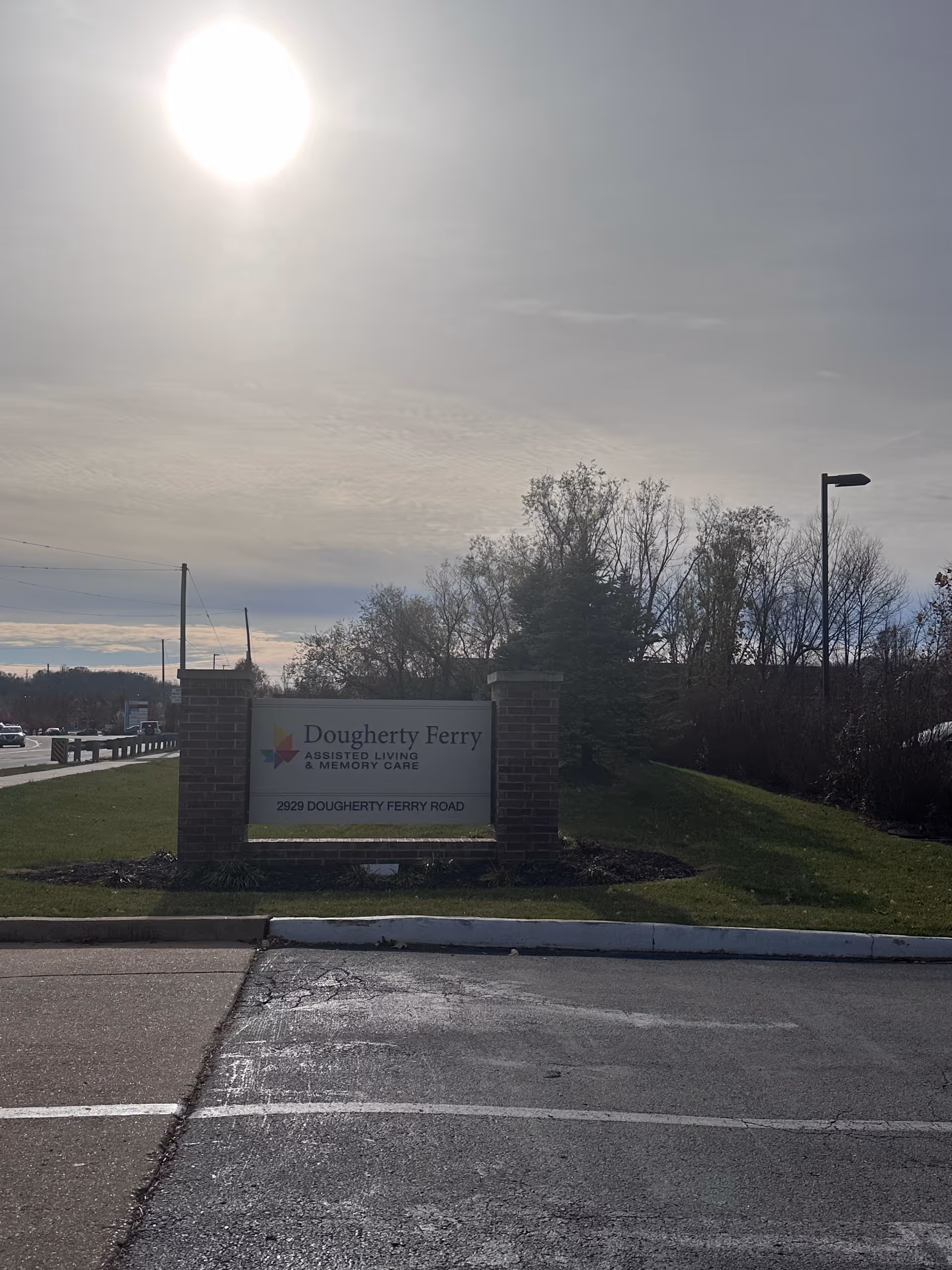 Outdoor view of a sign for Dougherty Ferry Assisted Living and Memory Care located at 2929 Dougherty Ferry Road. The sign is mounted between two brick pillars with a grassy area and trees in the background under a bright sun and cloudy sky.