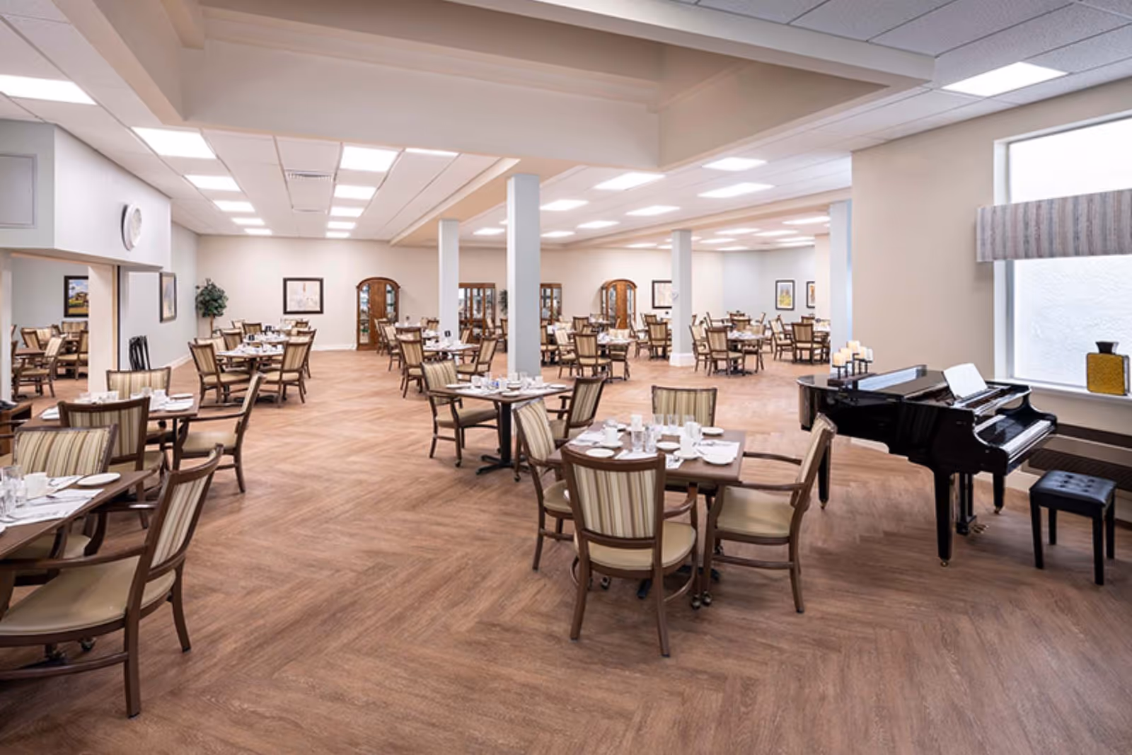 A spacious dining room with multiple wooden tables and chairs arranged neatly. Each table is set with white plates, cups, and silverware. The room has a light wood floor and white walls with framed artwork. There is a black grand piano with a stool near a large window on the right side of the room.