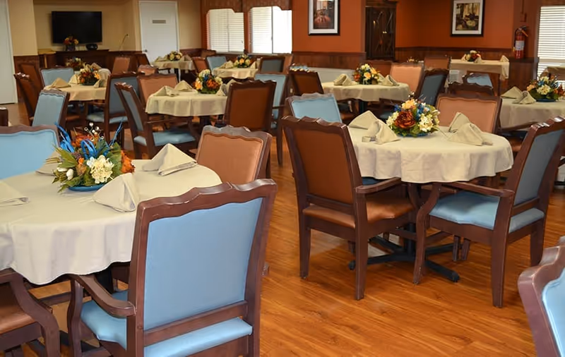 Dining room with round tables covered in tablecloths, floral centerpieces, and wooden chairs on a wood floor.