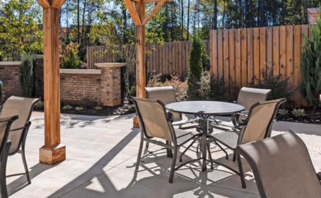 Sunlit outdoor patio with a round metal table and several mesh chairs under a wooden pergola beside a wood fence and landscaping.