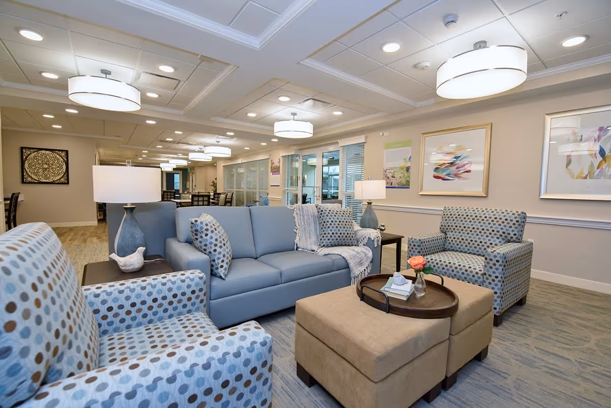 A well-lit living room area in a senior living facility featuring a light blue sofa with patterned pillows, two matching armchairs with a polka dot design, a beige ottoman with a tray holding a small vase with a pink flower and books, two table lamps, and framed artwork on the walls. In the background, there is a dining area with tables and chairs and large windows with blinds.