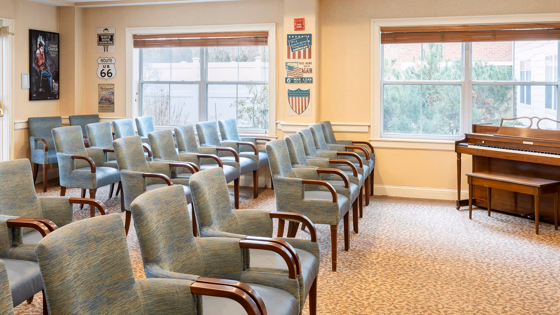 A room with multiple rows of blue upholstered chairs with wooden armrests arranged facing a piano near two large windows. The walls are decorated with vintage-style signs and a framed poster. The room has beige walls and patterned carpet flooring.