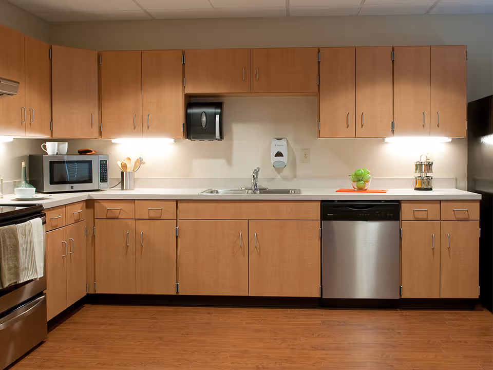 A clean and modern kitchen with light wood cabinets, a stainless steel dishwasher, a microwave, a sink with a faucet, and various kitchen utensils and items on the countertop including a bowl of green apples and a spice rack.