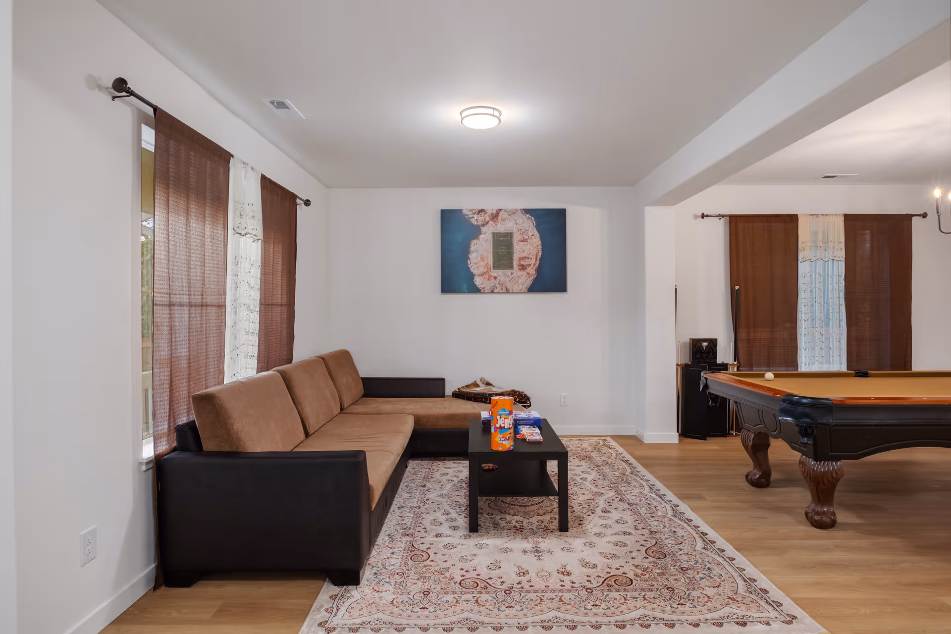 A living room area with a brown and black sectional sofa, a black coffee table with snacks on it, a patterned area rug, and a pool table in the adjacent space. The room has white walls, wooden floors, and windows with brown and white curtains. A framed artwork hangs on the wall above the sofa.