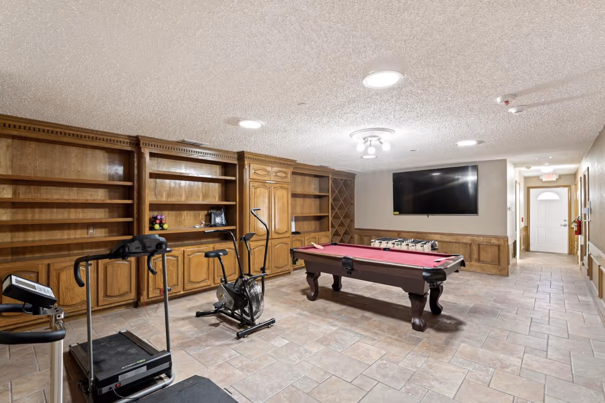A recreational room with a treadmill, exercise bike, pool table with a red felt top, and a foosball table. The room has wooden built-in shelves and cabinets along one wall, a large flat-screen TV mounted on another wall, tiled flooring, and ceiling lights.