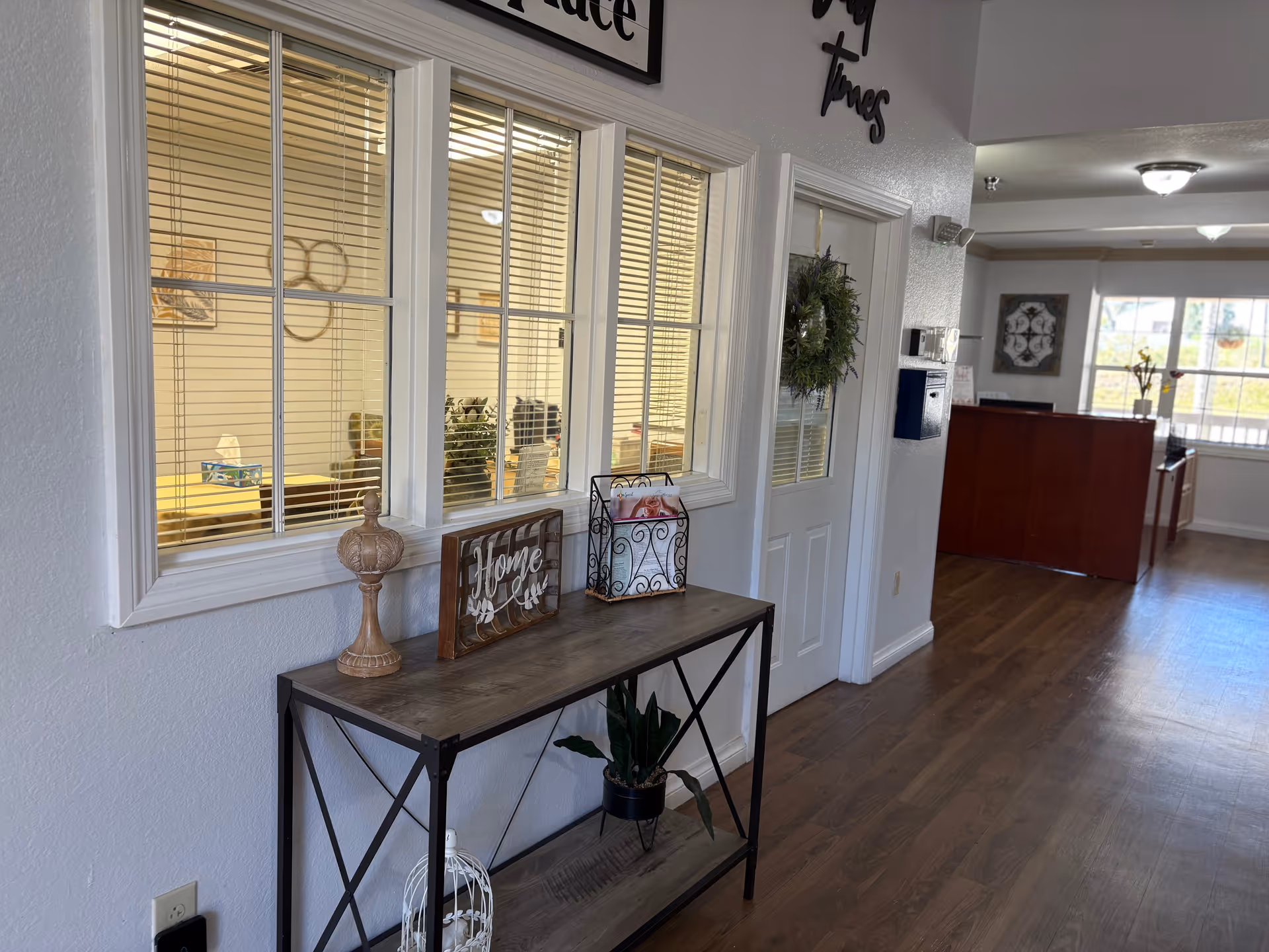 Interior hallway of a senior living facility with wooden flooring, a console table decorated with a plant, a decorative wooden piece, and a metal basket. There are windows with blinds on the left wall and a door with a wreath. In the background, a reception desk and large windows letting in natural light are visible.
