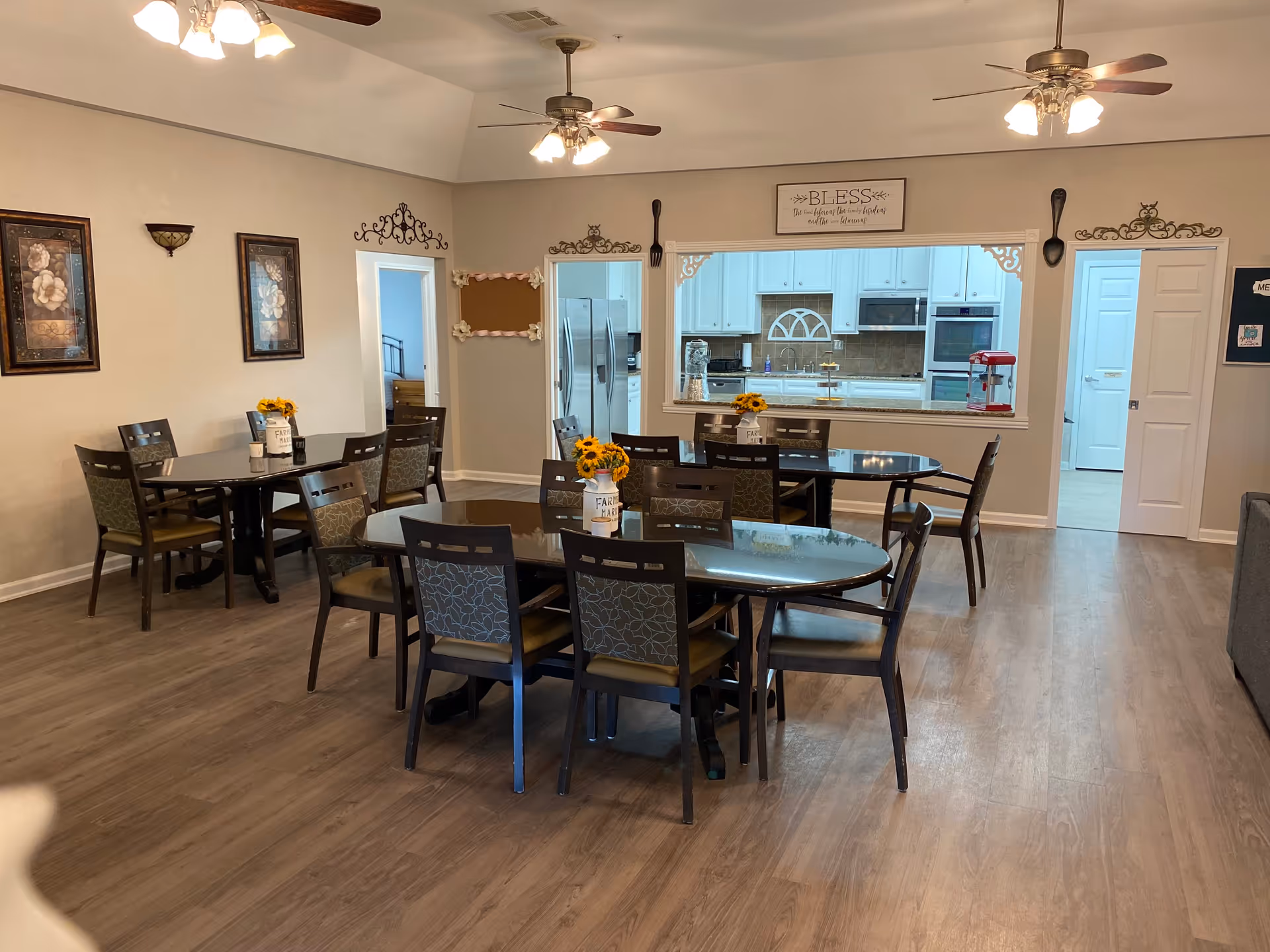 A dining room with three oval dark wood tables, each surrounded by six chairs with patterned backs and cushioned seats. Each table has a small vase with sunflowers. The room has light-colored walls with framed floral artwork and decorative wall accents. Ceiling fans with lights hang from the ceiling. A large open window connects the dining room to a white kitchen with stainless steel appliances and a popcorn machine on the counter.