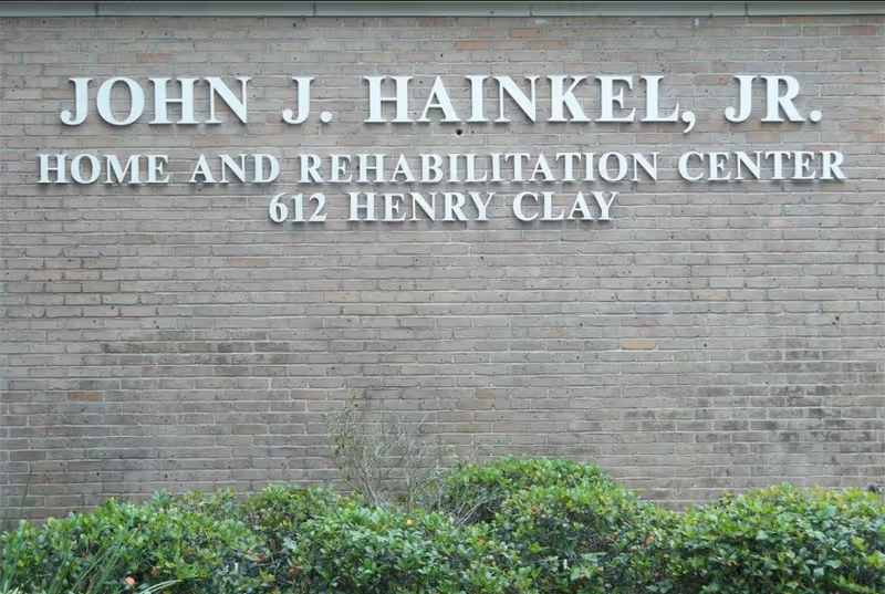 Exterior brick wall with white letters displaying the name 'John J. Hainkel, Jr. Home and Rehabilitation Center 612 Henry Clay' above green shrubbery.