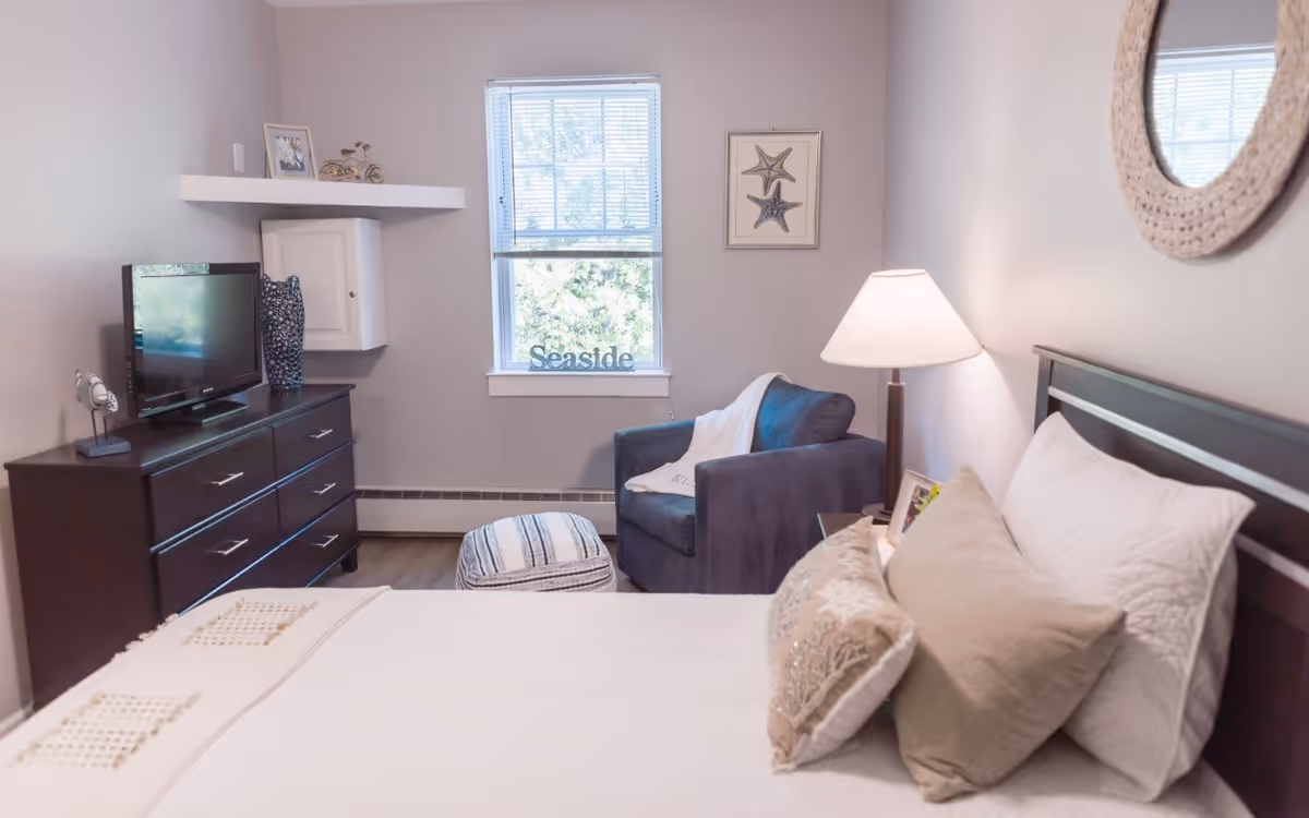A cozy bedroom with a neatly made bed featuring multiple pillows, a bedside table with a lamp, a comfortable armchair with a throw blanket, a window with blinds, a dresser with a TV and decorative items, and a round mirror on the wall.