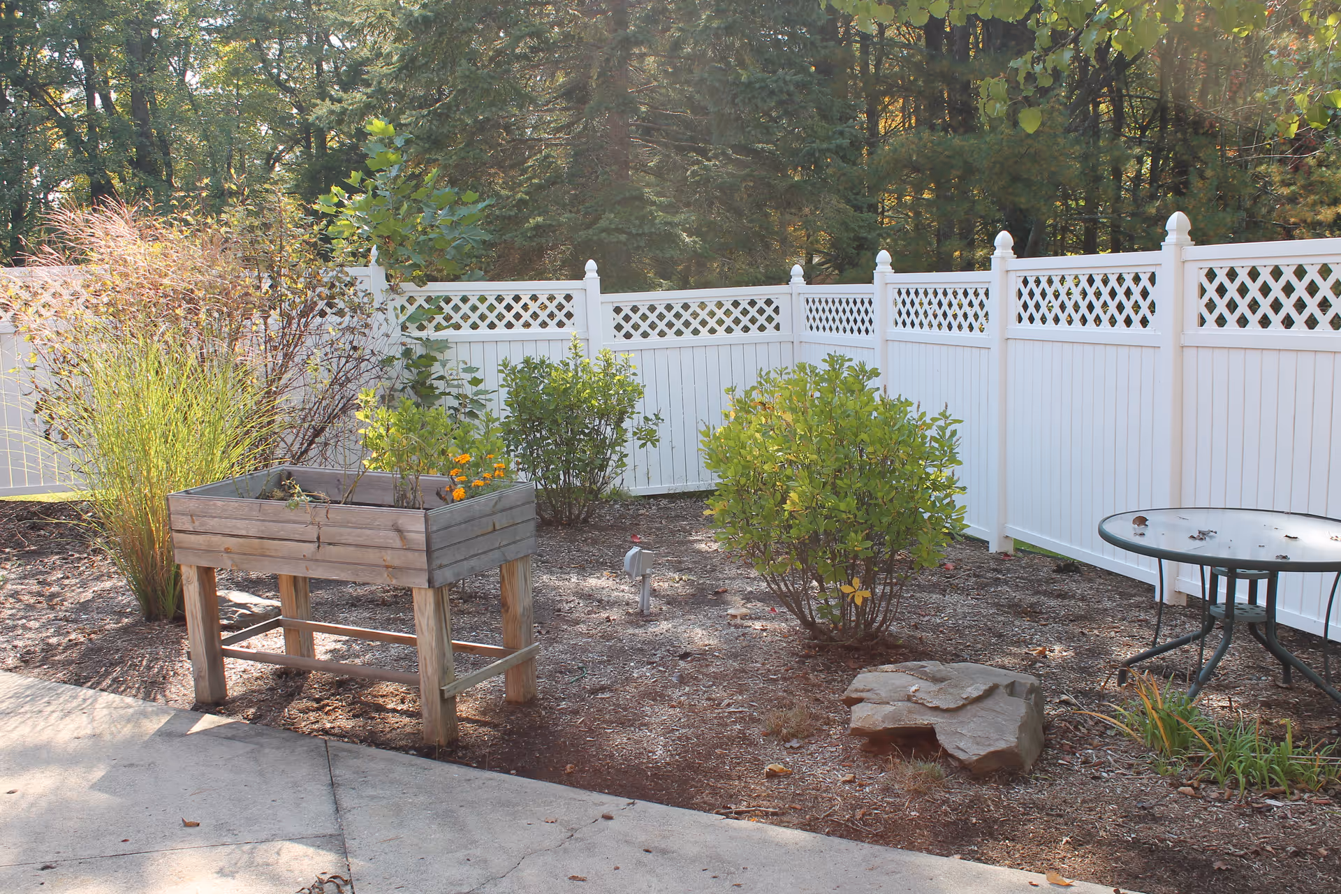 Outdoor garden area with a white lattice fence, a raised wooden planter box with some plants, green bushes, a round glass-top table, and a stone on the ground. Trees and greenery are visible in the background.