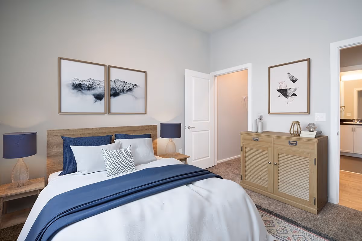A neatly made bed with white and blue bedding in a bedroom. There are two framed pictures of mountains above the wooden headboard. Two bedside tables with matching blue lamps flank the bed. To the right, there is a wooden cabinet with decorative items and a framed abstract artwork above it. Two open doors reveal a walk-in closet and a bathroom.