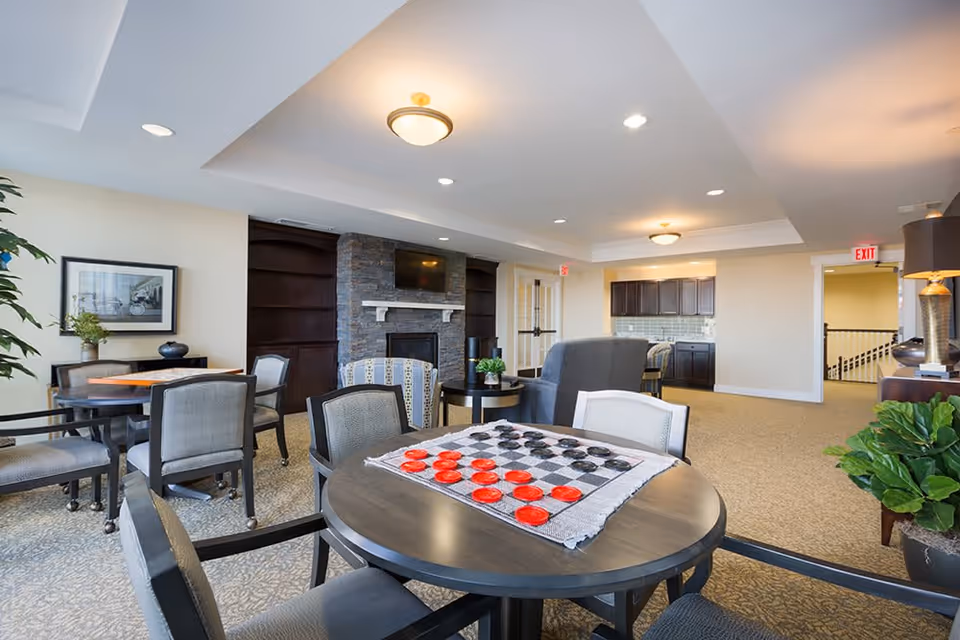 A bright and spacious common area in a senior living facility featuring a round table with a checkers game set up, several chairs, a stone fireplace with a mounted TV above it, additional seating areas with armchairs, a kitchenette with dark cabinets, and decorative plants and artwork on the walls.