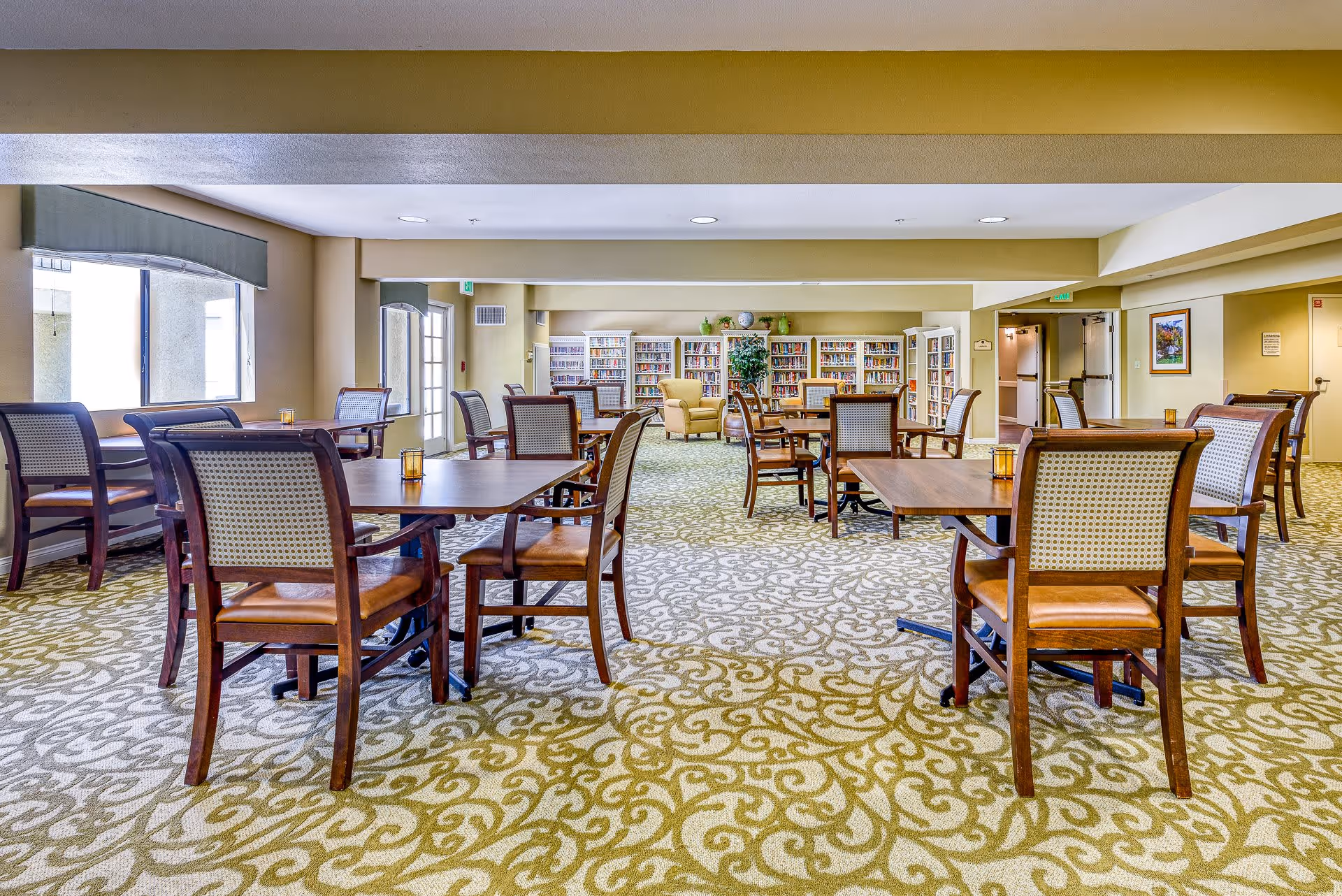 A spacious senior living common area with multiple wooden tables and chairs arranged on a patterned carpet. The room has large windows on the left side allowing natural light to enter. At the far end, there is a bookshelf filled with books and a comfortable armchair next to a potted plant. The walls are painted in a soft beige color, and the ceiling has recessed lighting.