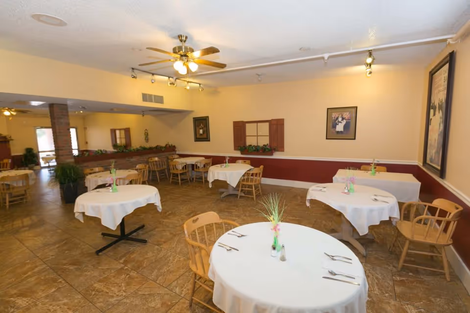 A dining room with several round and rectangular tables covered with white tablecloths. Each table has wooden chairs and small floral centerpieces. The room has beige walls with a maroon lower section, framed pictures on the walls, ceiling fans with lights, and tiled flooring.