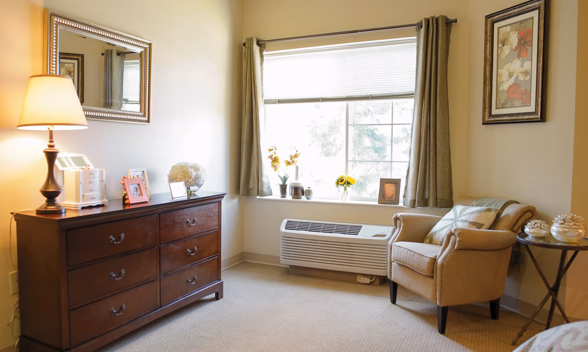 Sunny sitting area with a wooden dresser and lamp on the left and an upholstered armchair beside a window on the right.