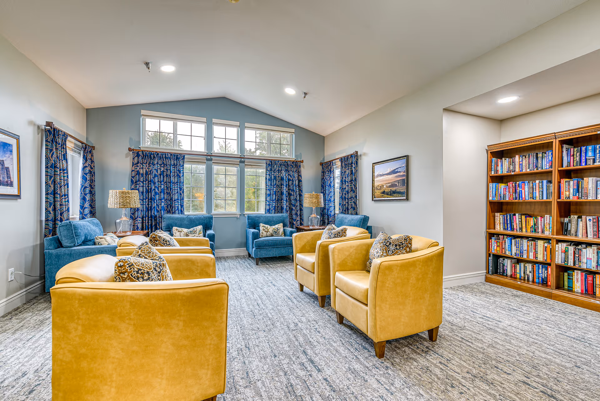 Communal lounge with yellow and blue armchairs arranged facing each other, large windows with patterned curtains, lamps, and a bookshelf.