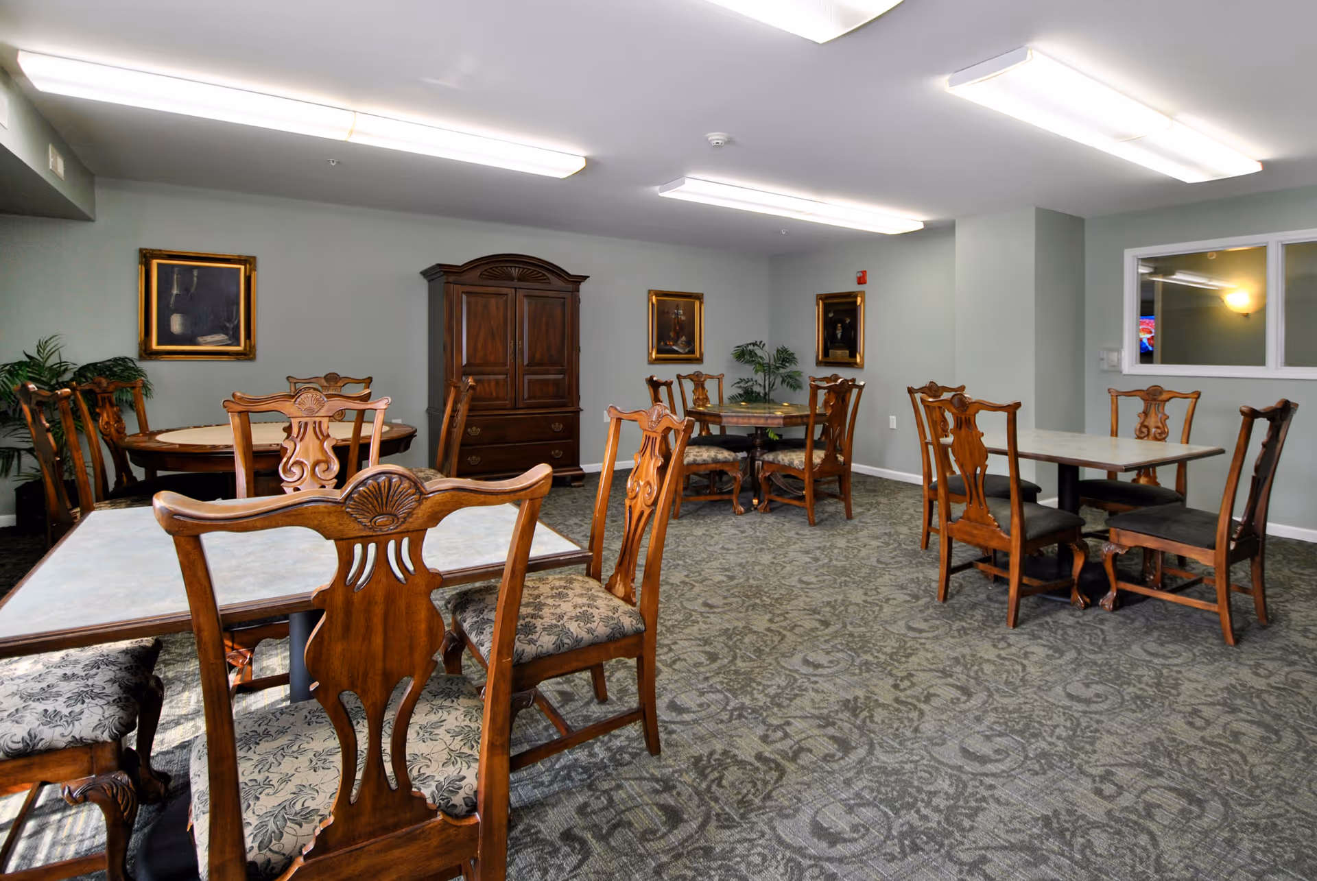 Communal dining room with multiple wooden tables and ornate chairs, carpeted floor, and framed artwork on the walls.