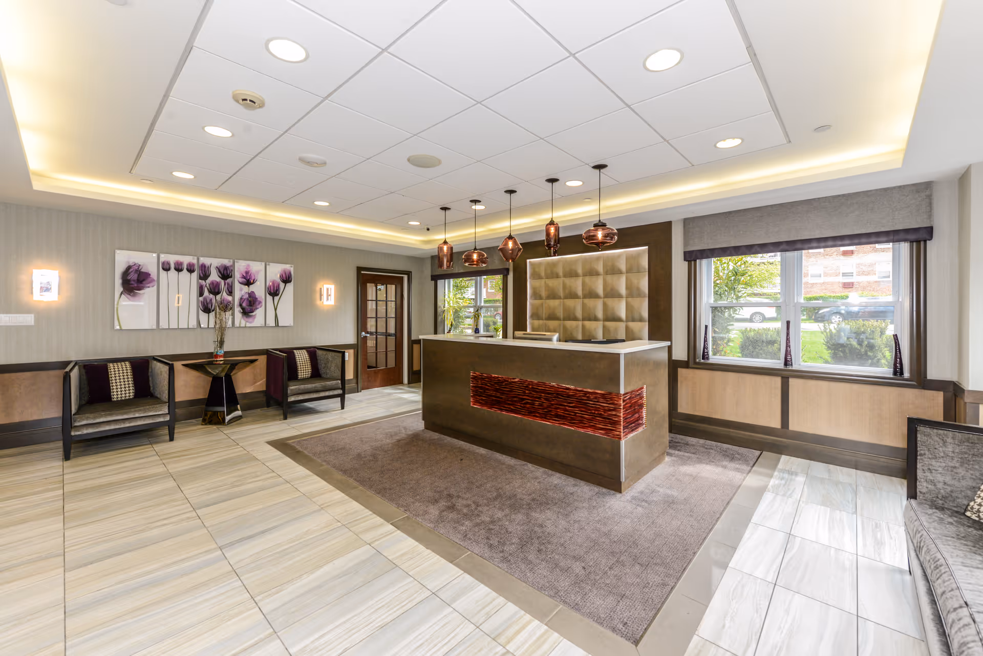 A modern and well-lit reception area with a central desk featuring a textured front panel and pendant lights hanging above. The room has tiled floors, a large window with a view of greenery outside, and seating areas with chairs and a small table. Wall art with purple floral designs decorates one wall.
