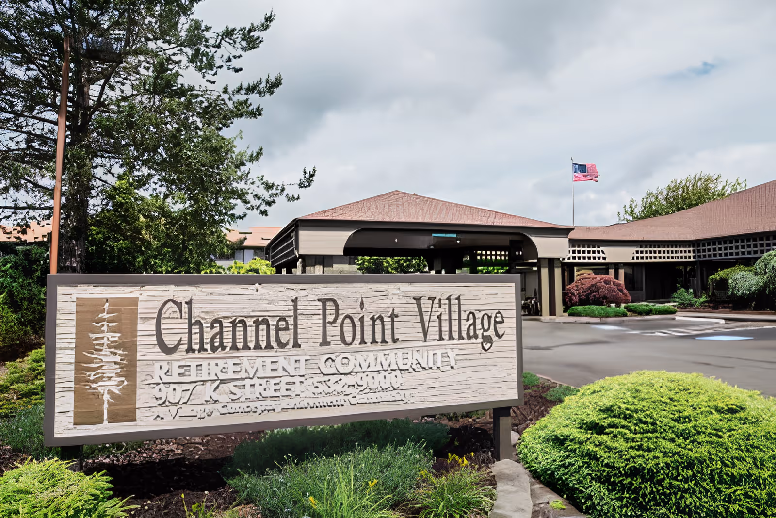 Entrance sign for Channel Point Village retirement community with a building and flagpole in the background surrounded by greenery and bushes under a cloudy sky.